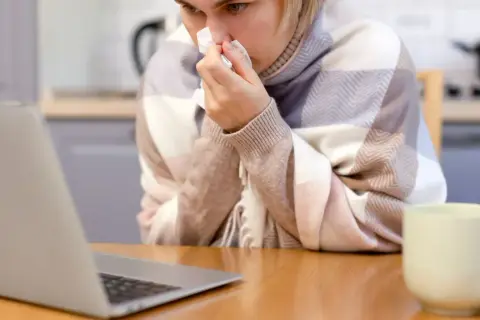 Kobieta w kratę, z chusteczką w dłoni, korzysta z laptopa. Może to być polskie zwolnienie lekarskie w Danii, gdy czuje się źle.