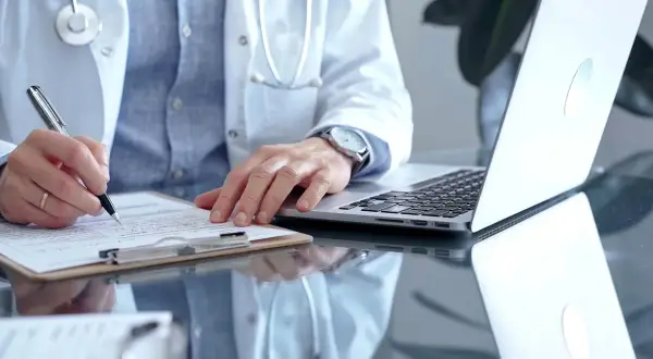 Lekarz rodzinny wypisuje receptę na depresję. Na biurku leży laptop i dokumenty.
