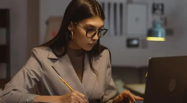 Kobieta w okularach pracuje przy laptopie, notując coś w zeszycie. Zastanawia się, ile maksymalnie nadgodzin w miesiącu może przepracować.