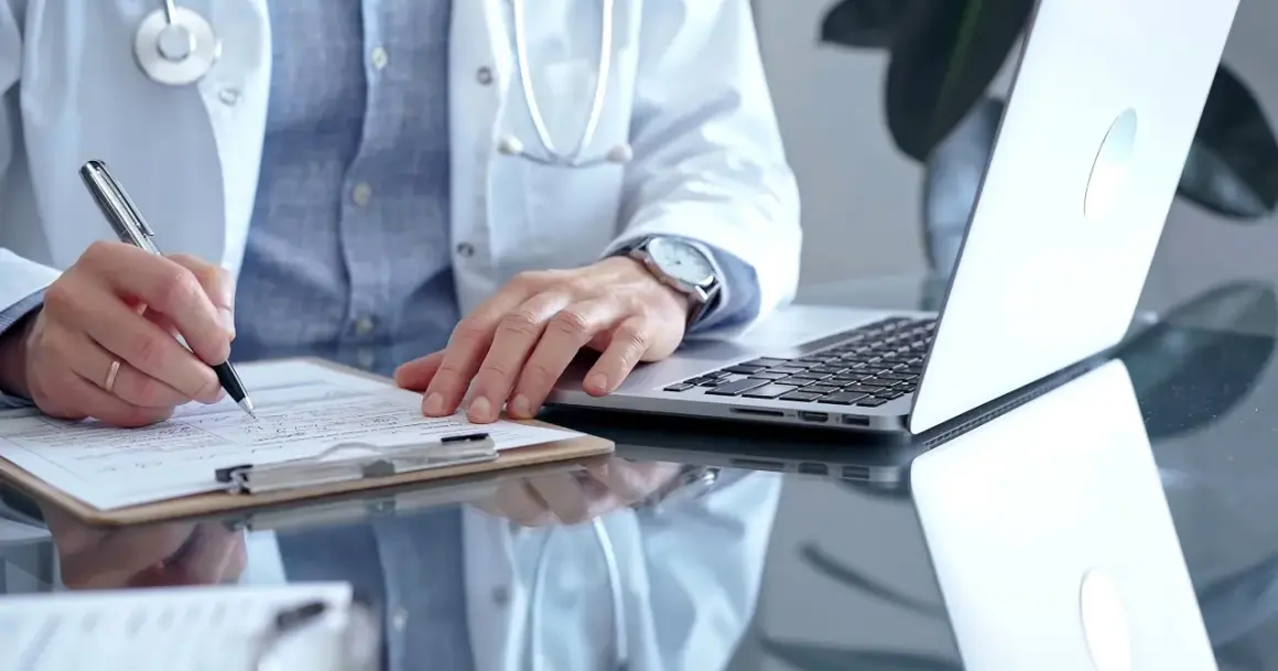 Lekarz rodzinny wypisuje receptę na depresję. Na biurku leży laptop i dokumenty.