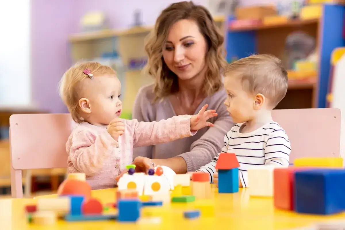 Nauczycielka i dwoje dzieci bawią się klockami przy żółtym stole.